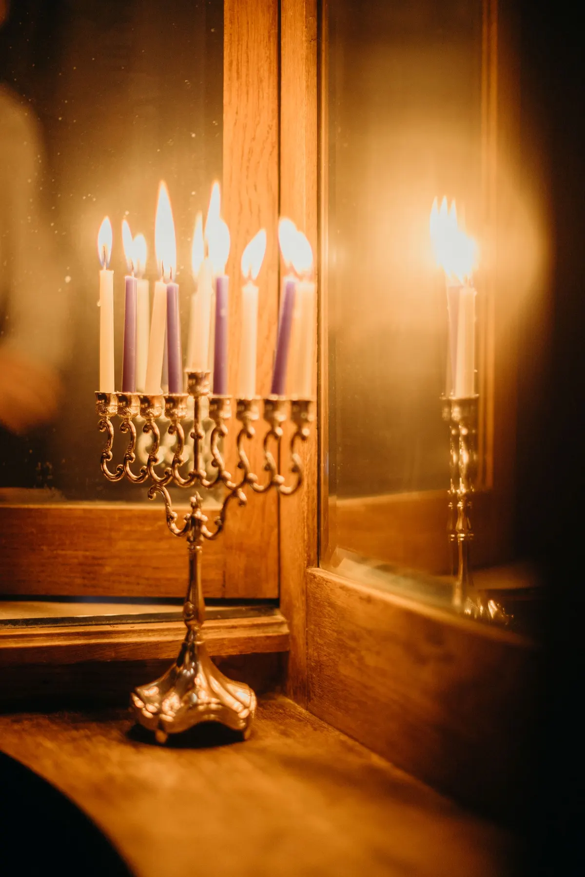 Gold menorah with nine lit ivory and purple candles on a wooden windowsill reflecting in the glass