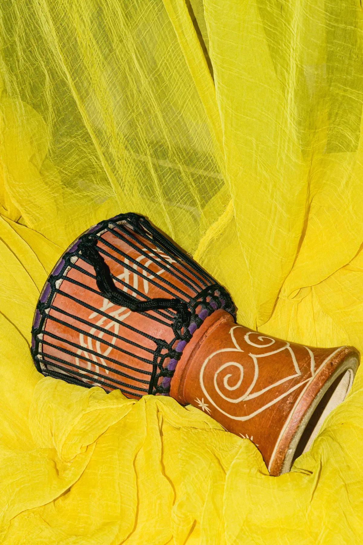 Carved orange-brown djembe drum with white swirls and black rope lacing lying on yellow fabric