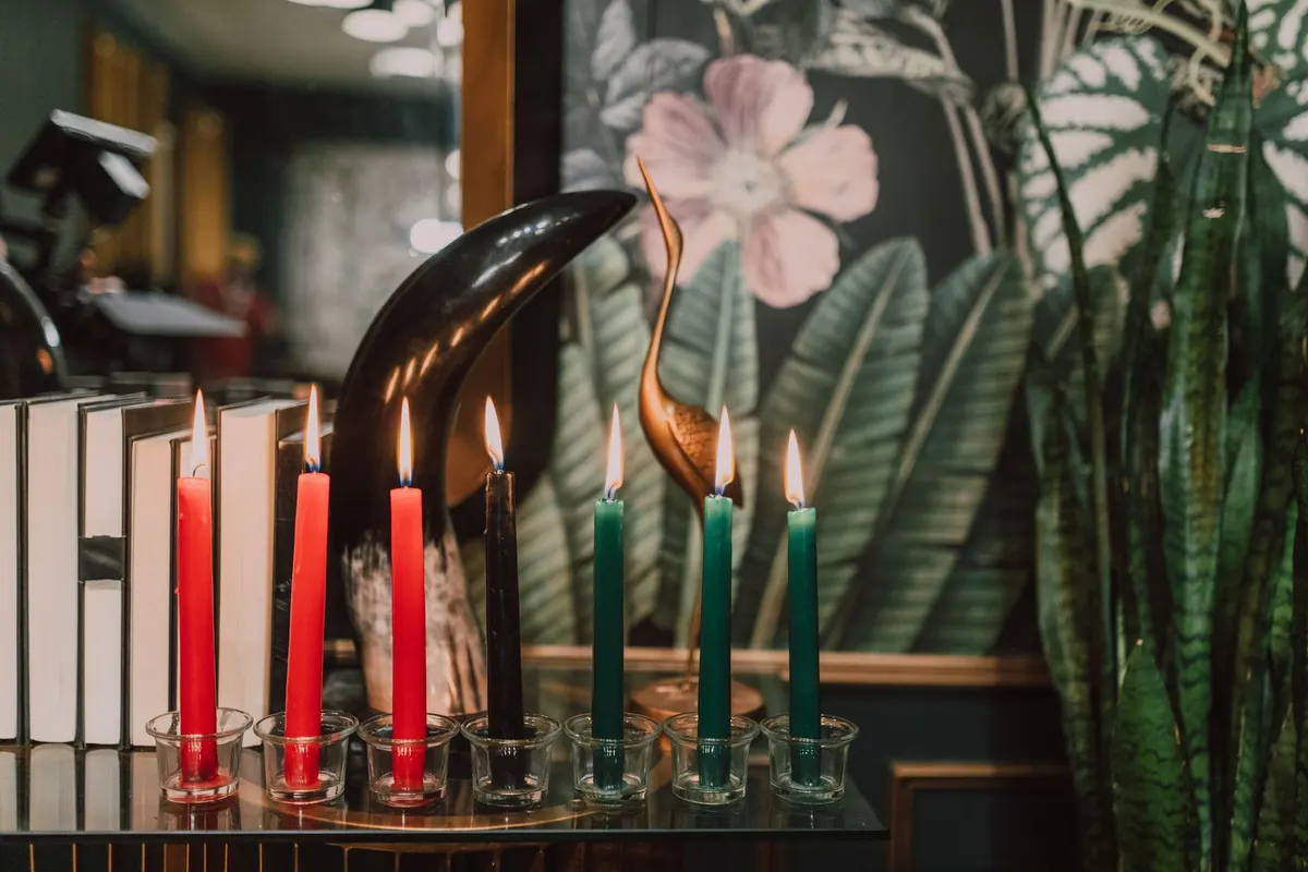Seven lit candles in clear glass cups on a glass console beside upright books and a brass bird sculpture
