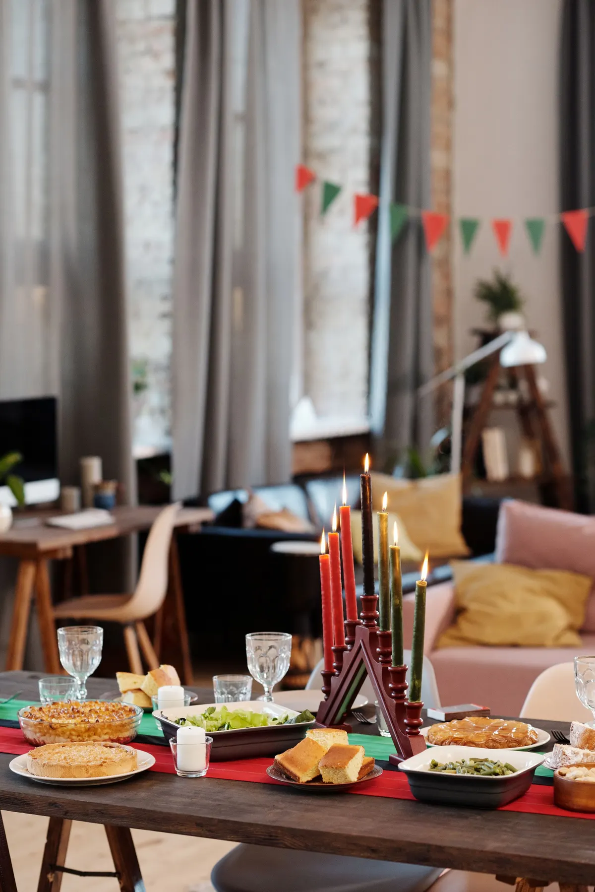 Feast table with a lit kinara, casseroles, cornbread, salad and stemmed glasses on a red runner