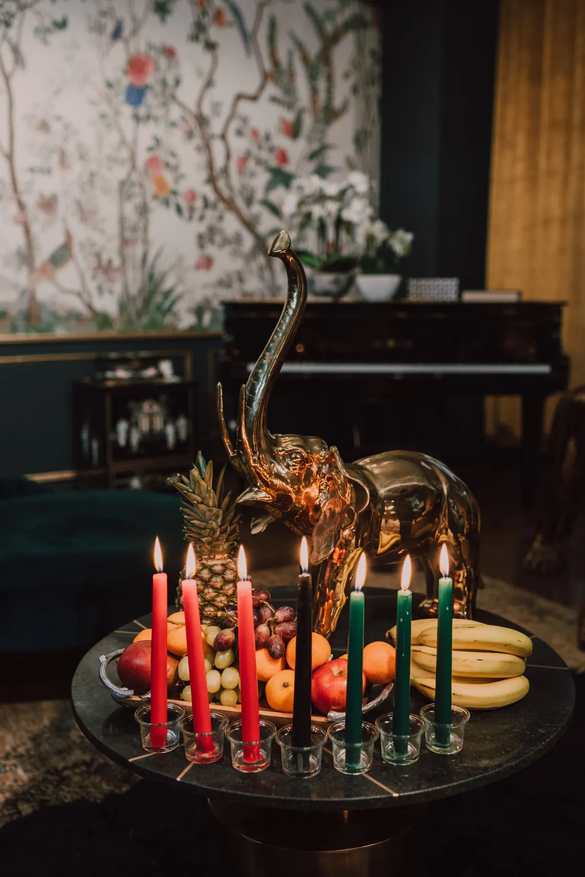 Seven lit red, black and green candles in glass cups before a silver tray of pineapple, grapes and oranges