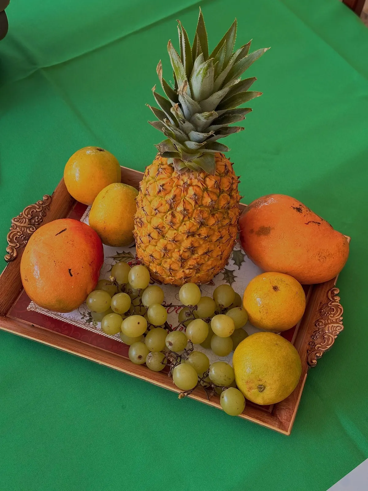 Wooden tray with brass handles holding a pineapple, two mangoes, four citrus fruits and green grapes