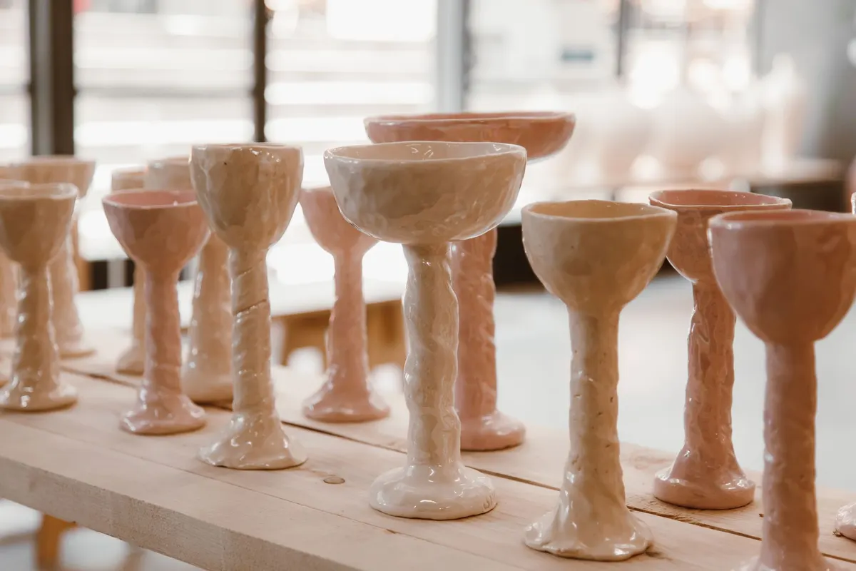 Rows of handmade glazed ceramic goblets in cream and pale pink standing on a pine bench