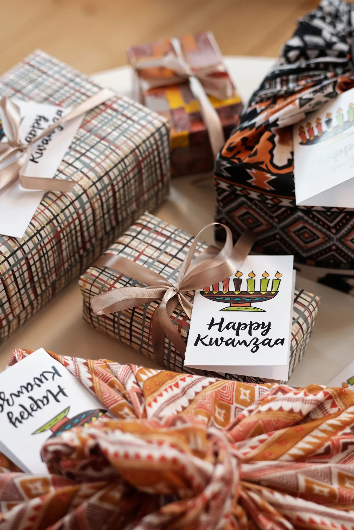 Five gifts wrapped in plaid paper and patterned African fabric beside white Happy Kwanzaa greeting cards