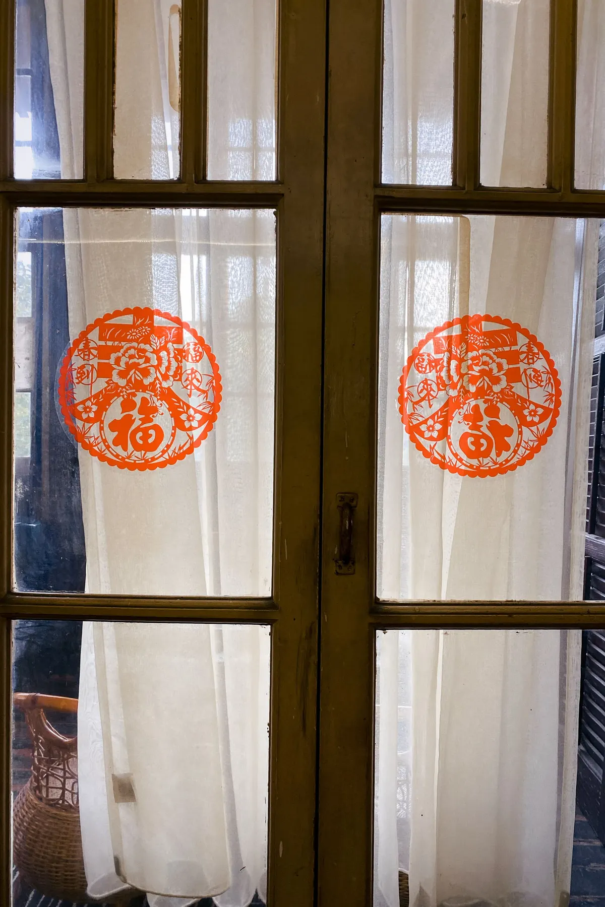 Two round orange-red floral paper cuttings on olive-framed glass doors with white curtains behind