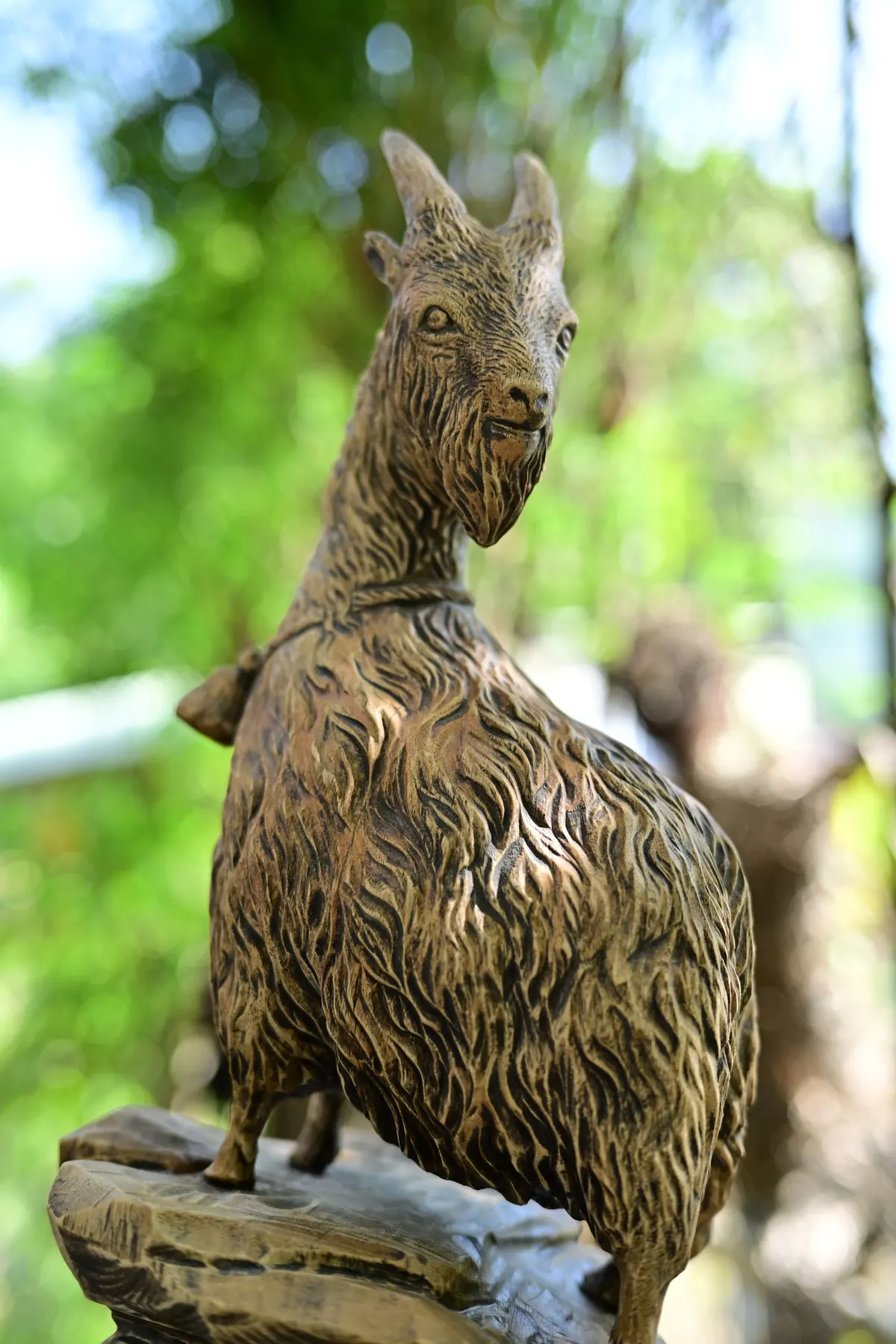 Carved brown wooden goat with curved horns and a small bell standing on a carved base