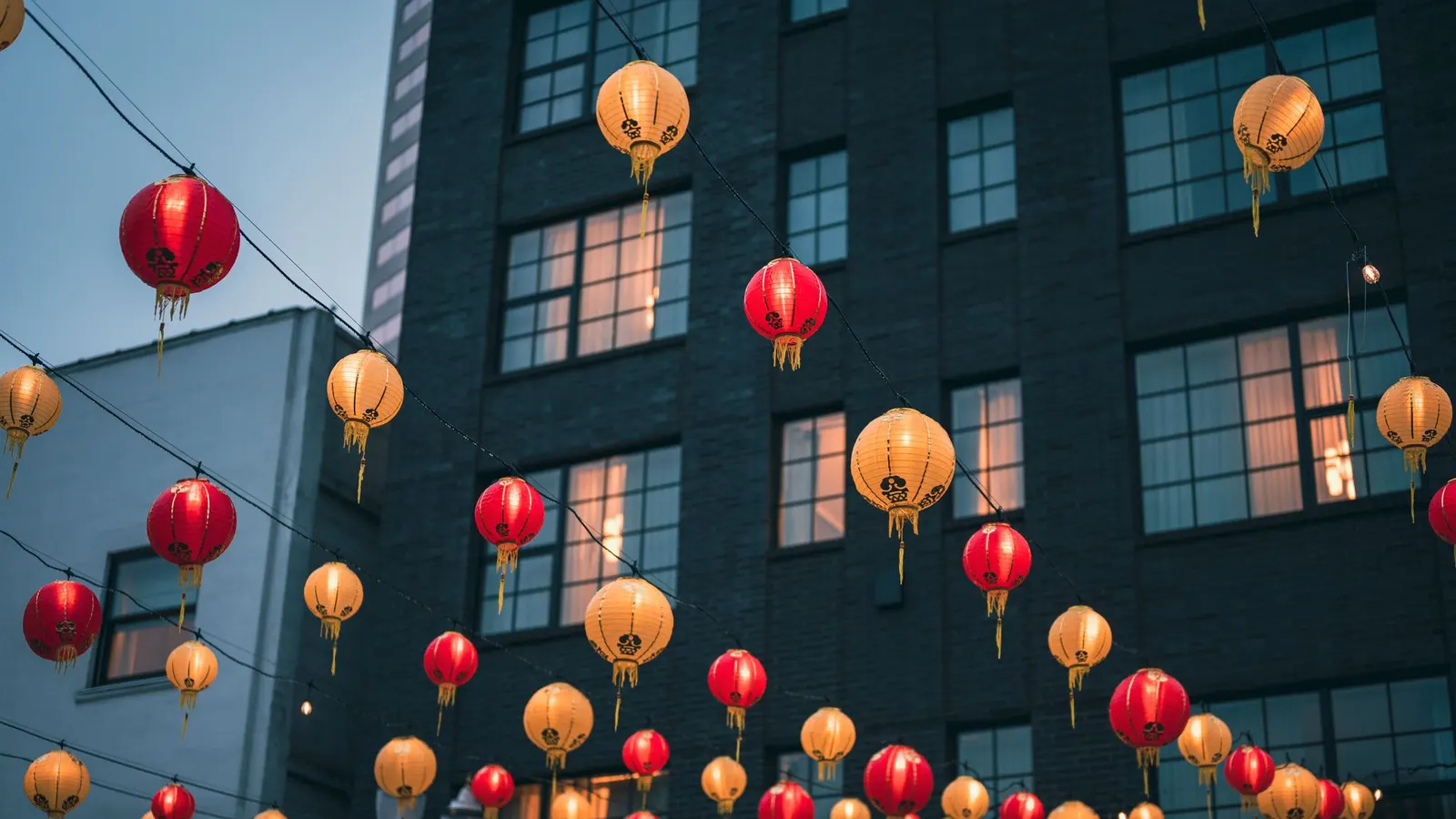 Rows of pale orange and red lanterns strung on wires across a street between gray city buildings