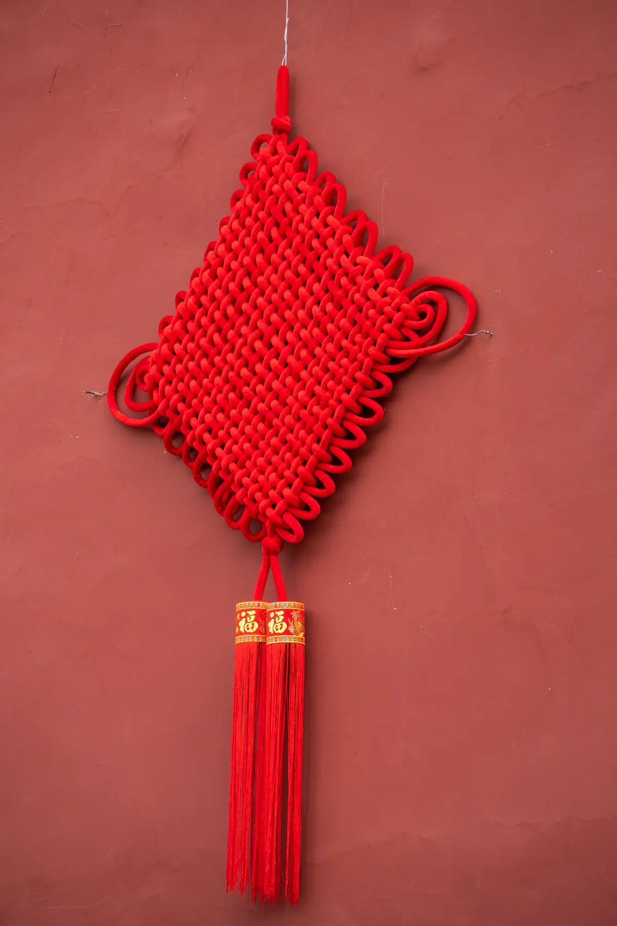 Red diamond-shaped woven Chinese knot with two long red tassels hanging against a reddish wall