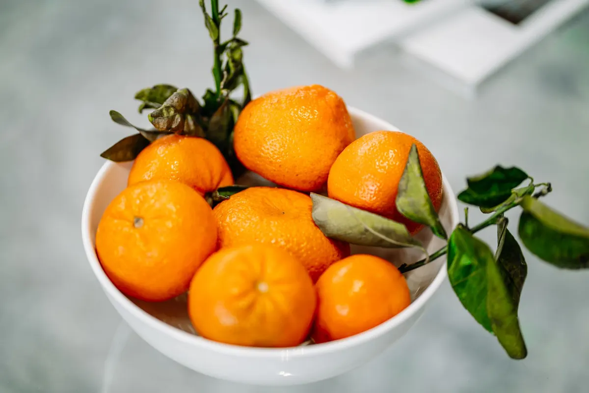 White bowl piled with mandarin oranges, some with green leaves still attached, on a gray surface