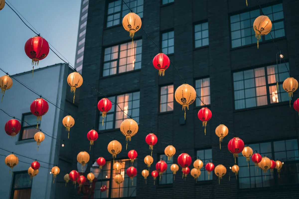 Rows of pale orange and red lanterns strung on wires across a street between gray city buildings