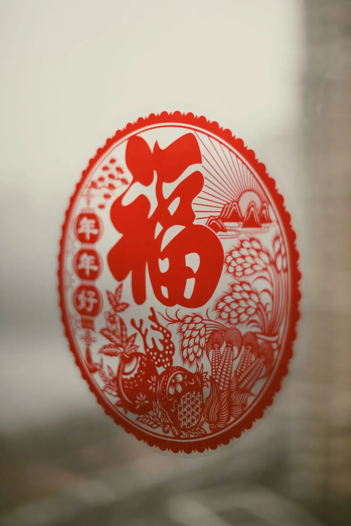 Oval red paper cutting with a large fu character, a rooster, corn and wheat stuck to window glass