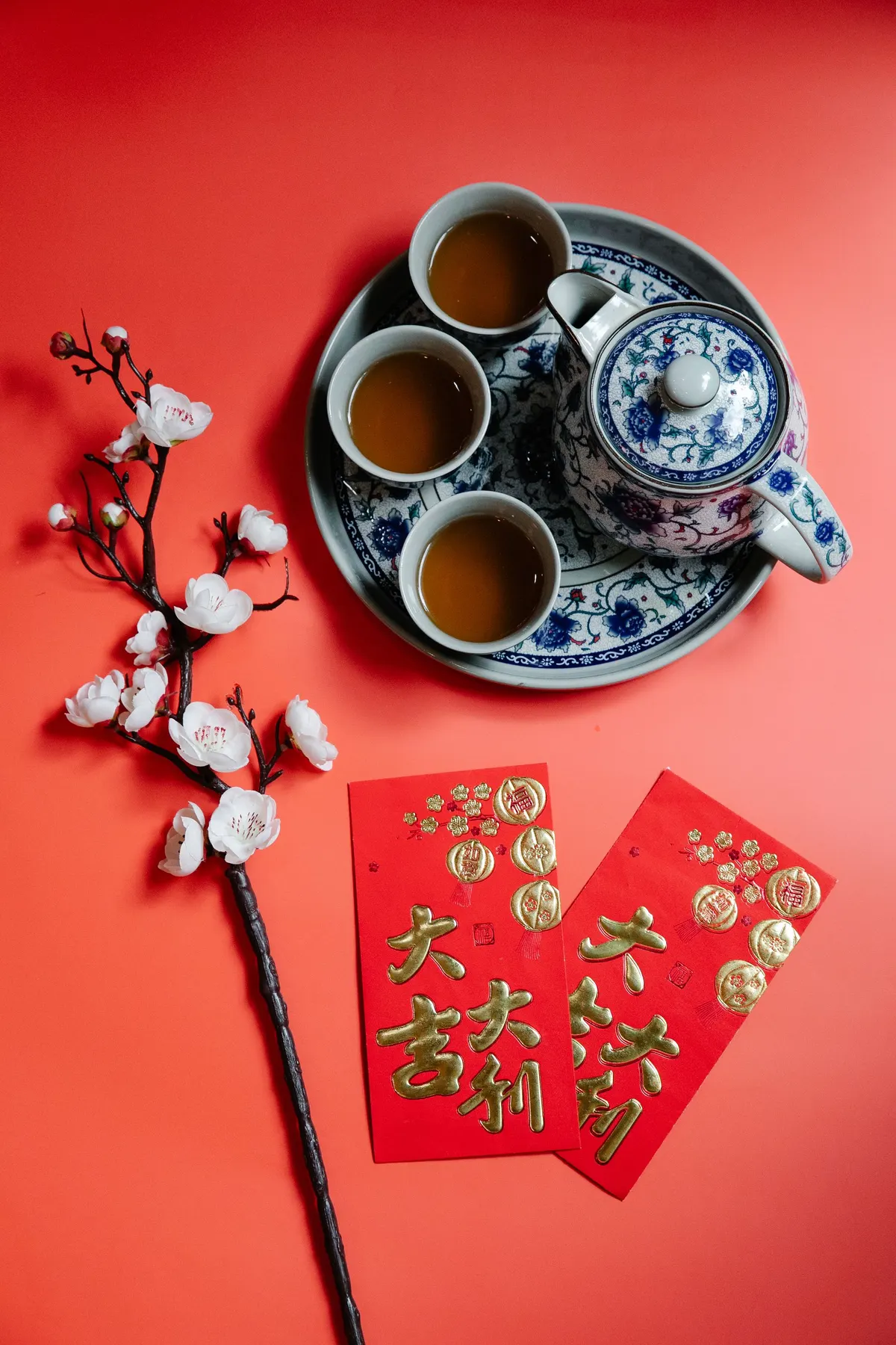 Two red and gold envelopes beside a blue and white teapot, three cups and a blossom branch