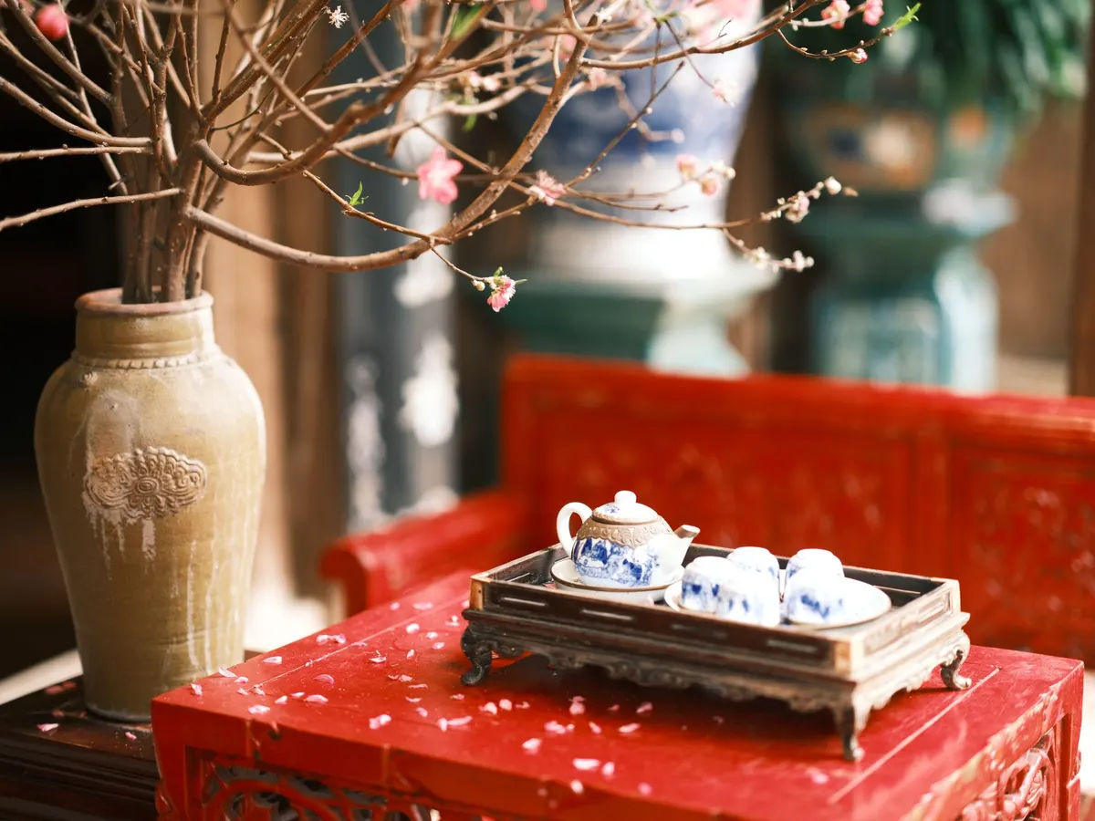 Blue and white teapot and four small cups on a dark tray beside a tall vase of pink blossoms