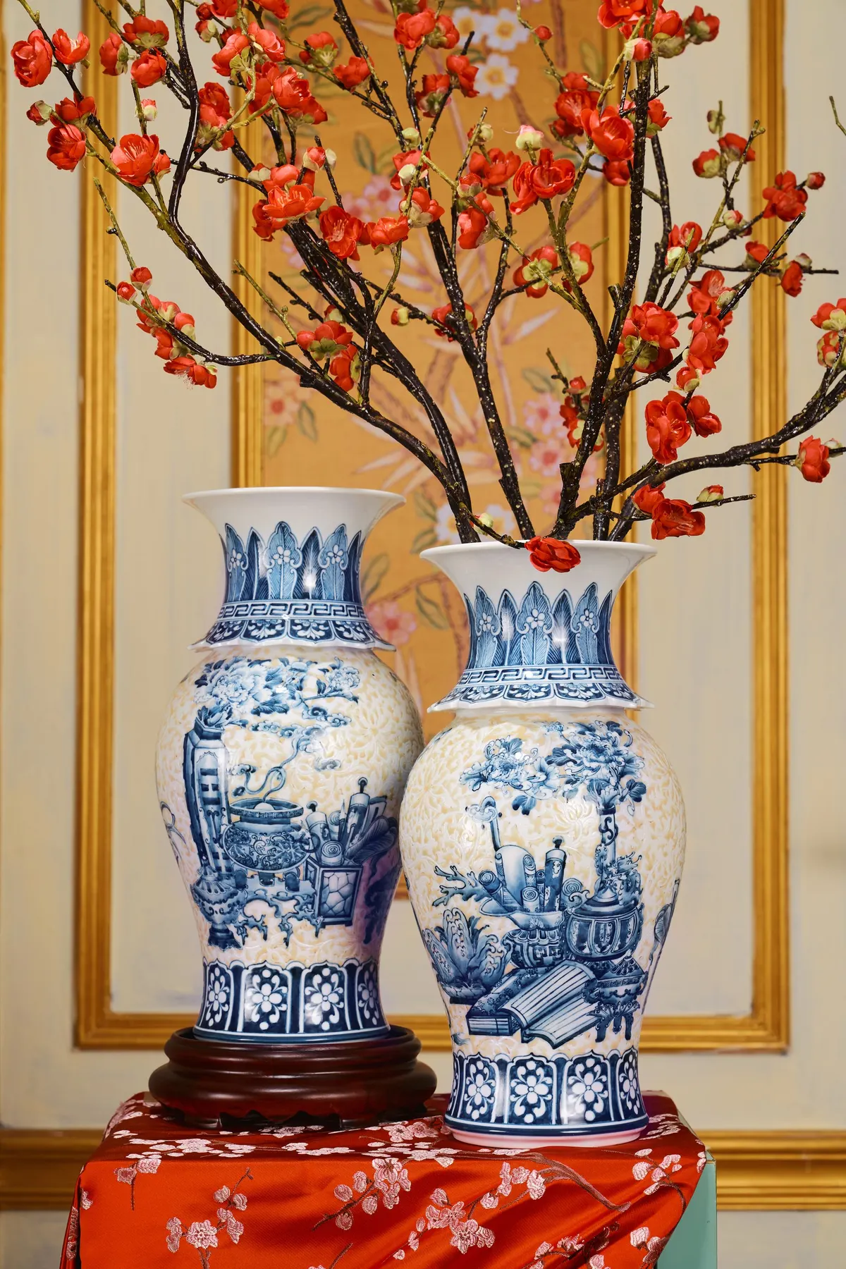 Two blue and white vases on red brocade, one on a dark wooden stand, one holding red blossom branches