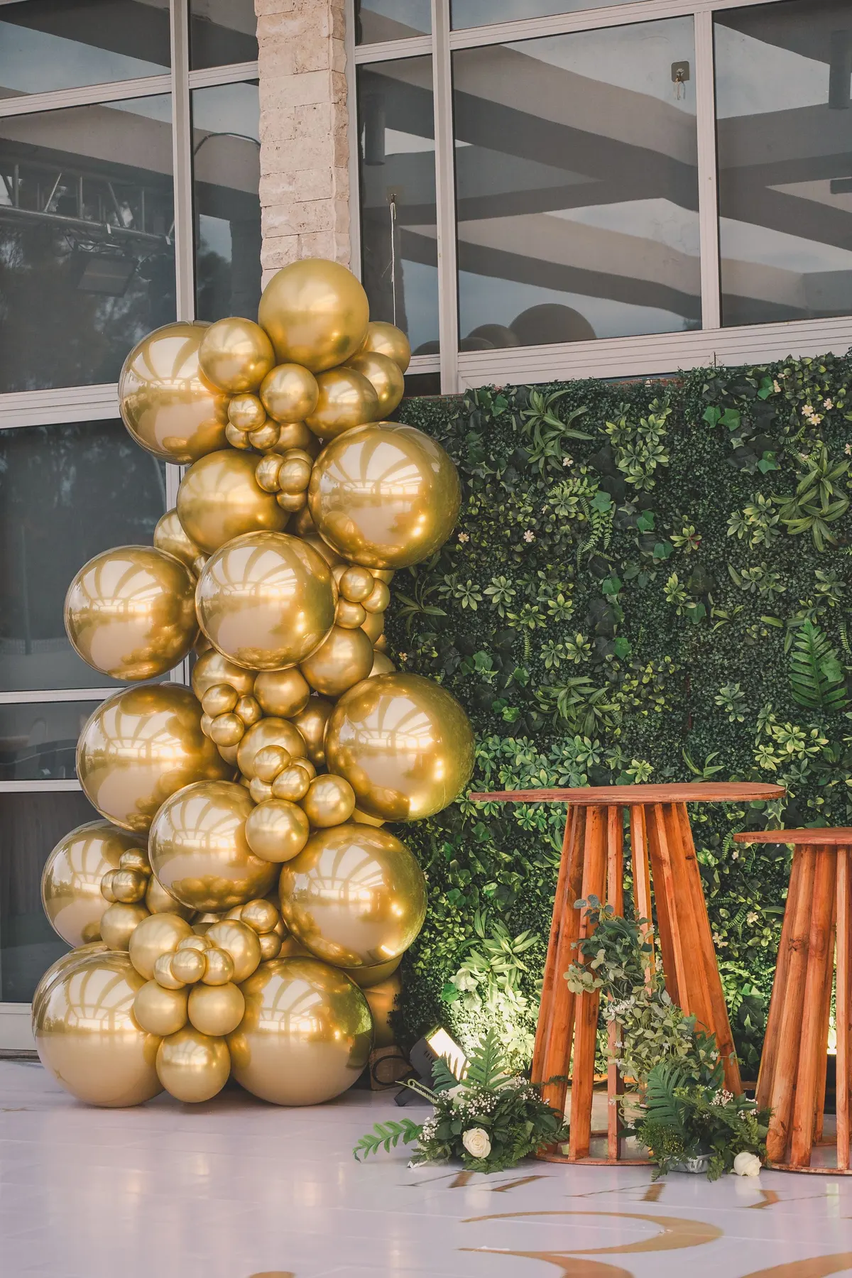 Tall column of gold balloons beside a green foliage wall with two wooden pedestal tables and white flowers