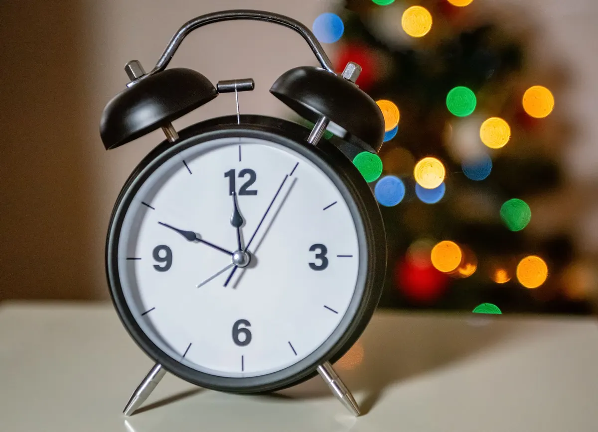 Black twin-bell alarm clock with a white face on a pale table in front of colorful blurred lights