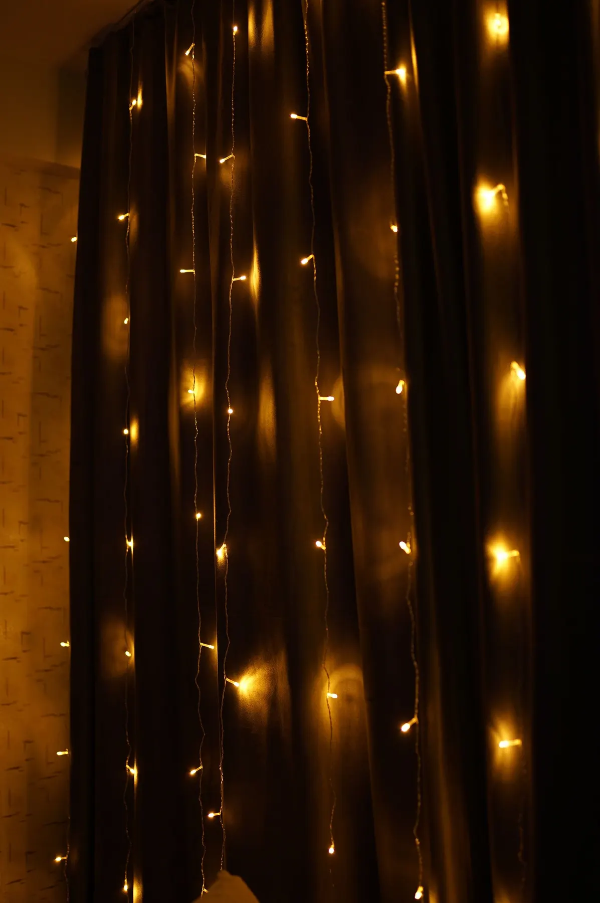 Vertical strands of warm yellow fairy lights hanging over a dark pleated curtain next to a beige wall