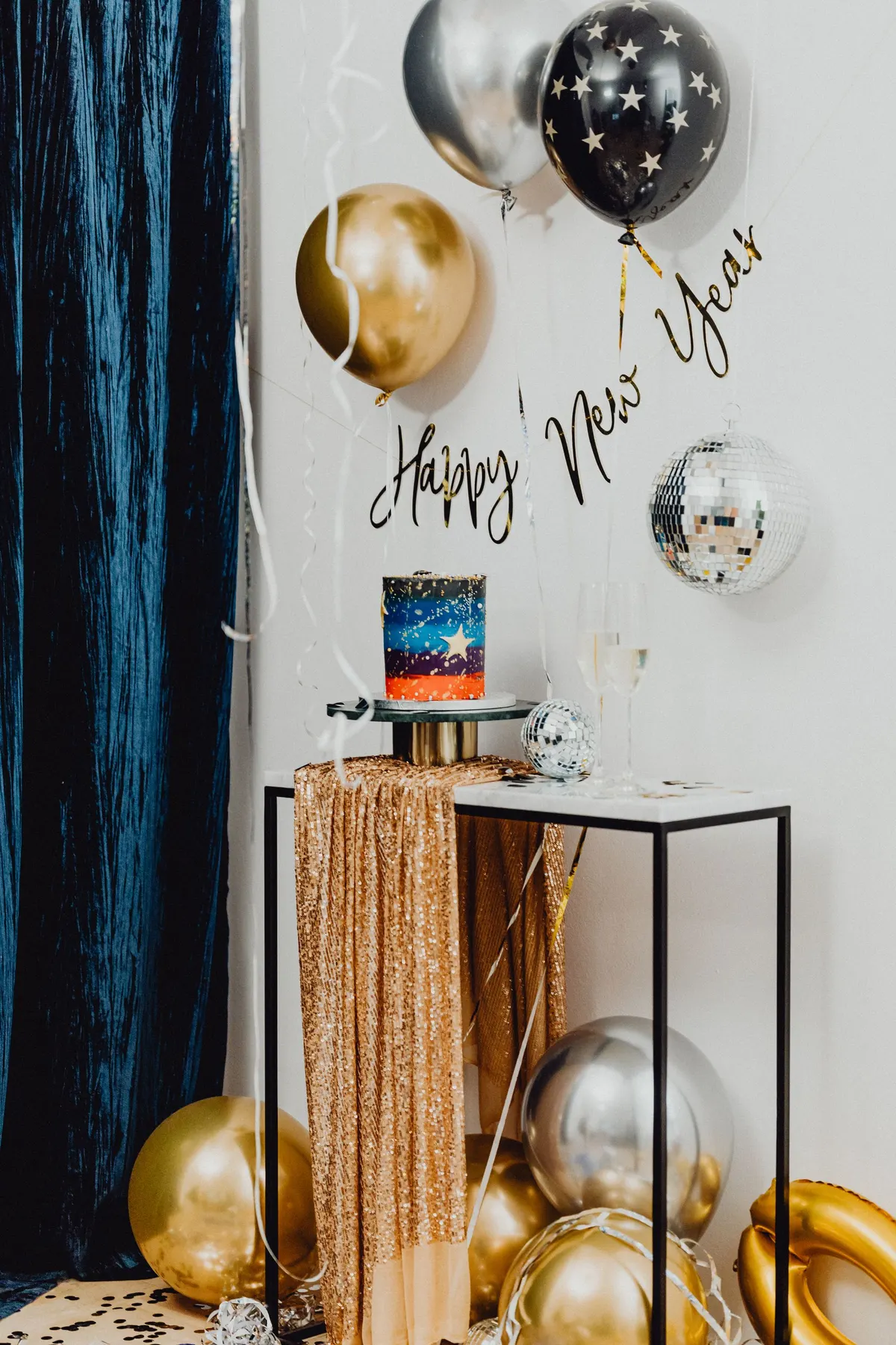 Dessert table with a layered cake on a stand, gold sequin drape, mirror balls and gold, silver and black balloons