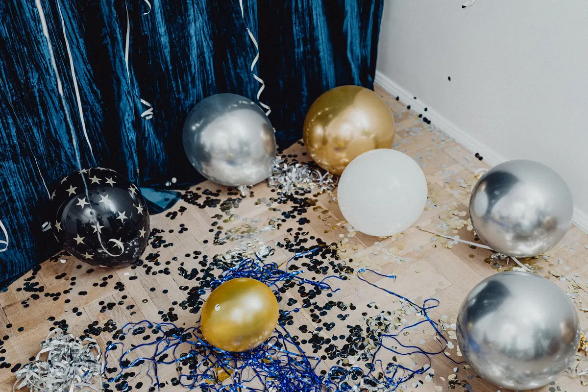 Seven gold, silver, white and black star balloons on a wood floor with confetti and blue ribbons