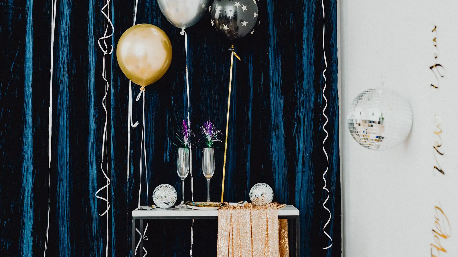 Gold, silver and black star balloons beside a table with flutes and mirror balls against a dark blue curtain