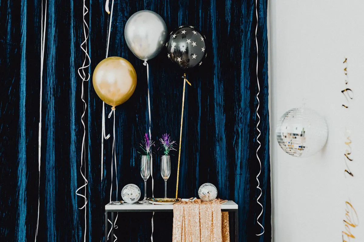 Gold, silver and black star balloons beside a table with flutes and mirror balls against a dark blue curtain