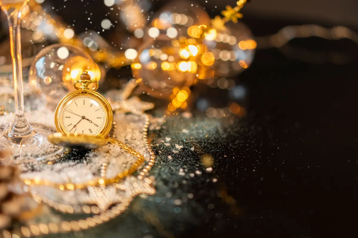 Open gold pocket watch with its chain beside a stemmed glass, clear baubles and gold strands on a dark tabletop