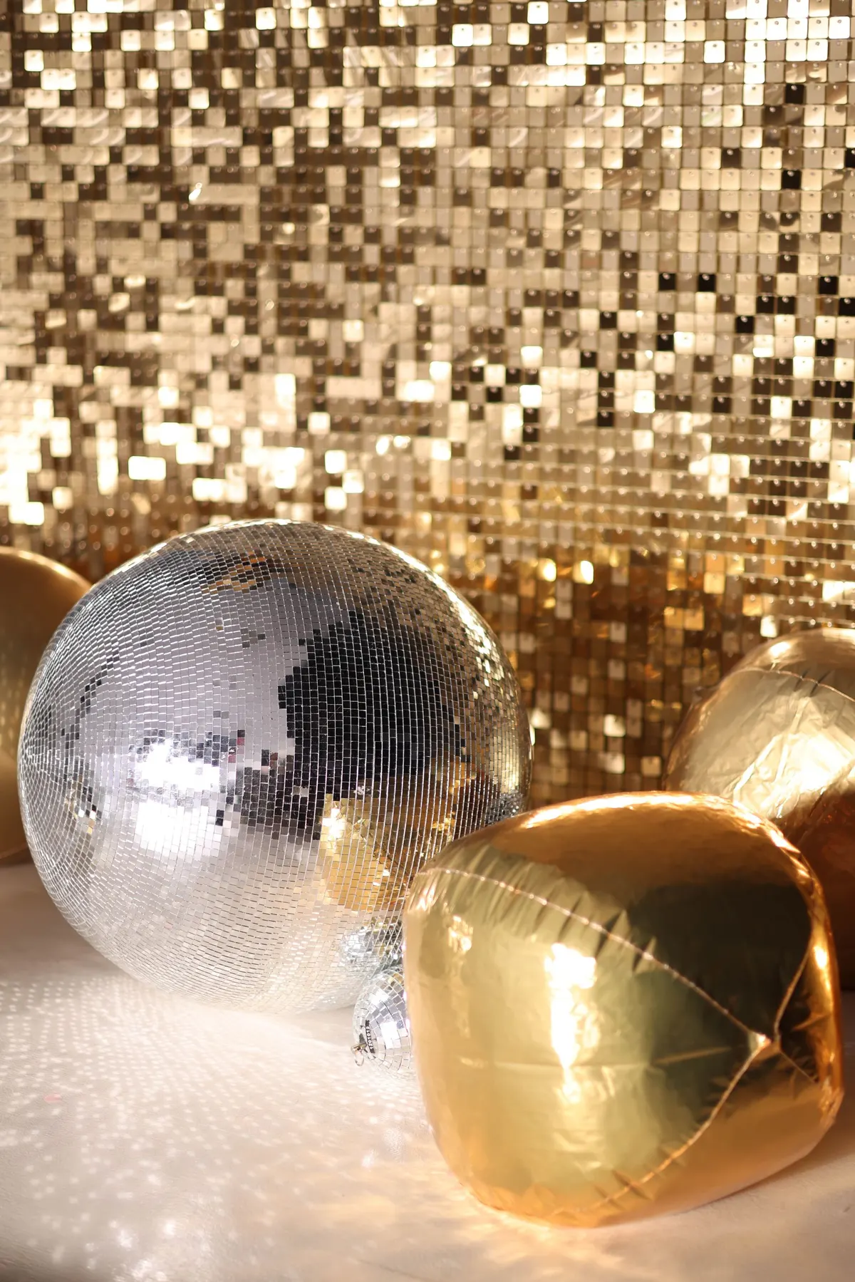 Large and small mirror balls with three gold balloons in front of a gold square sequin backdrop