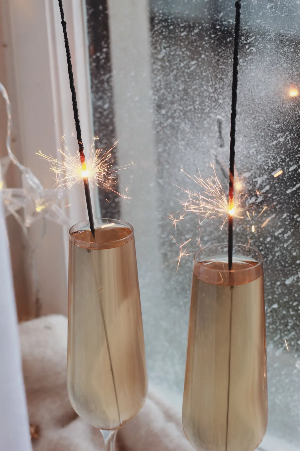 Two champagne flutes with lit black sparklers standing beside a frosted white window frame