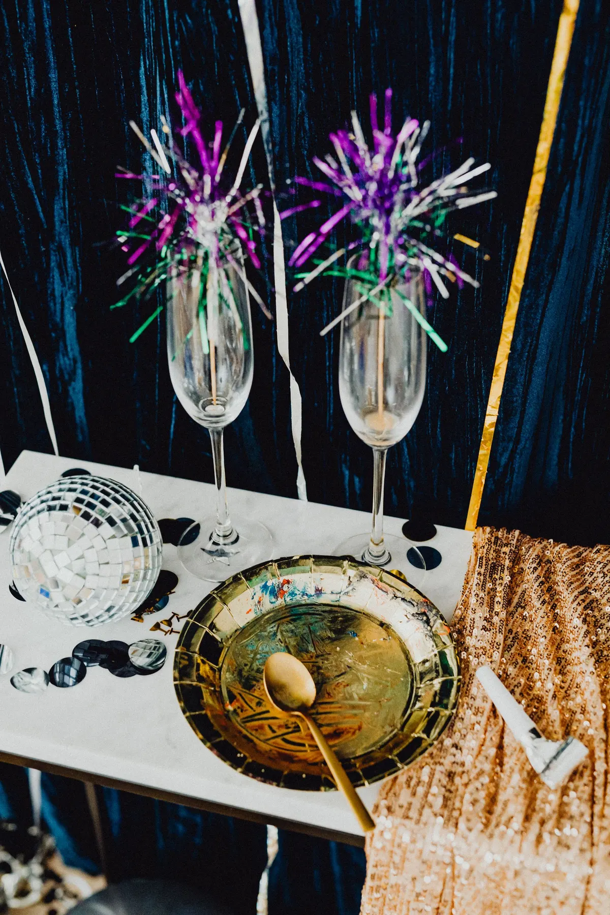 White party table with two tinsel-trimmed flutes, a mirror ball, gold plate, party blower and gold sequin fabric