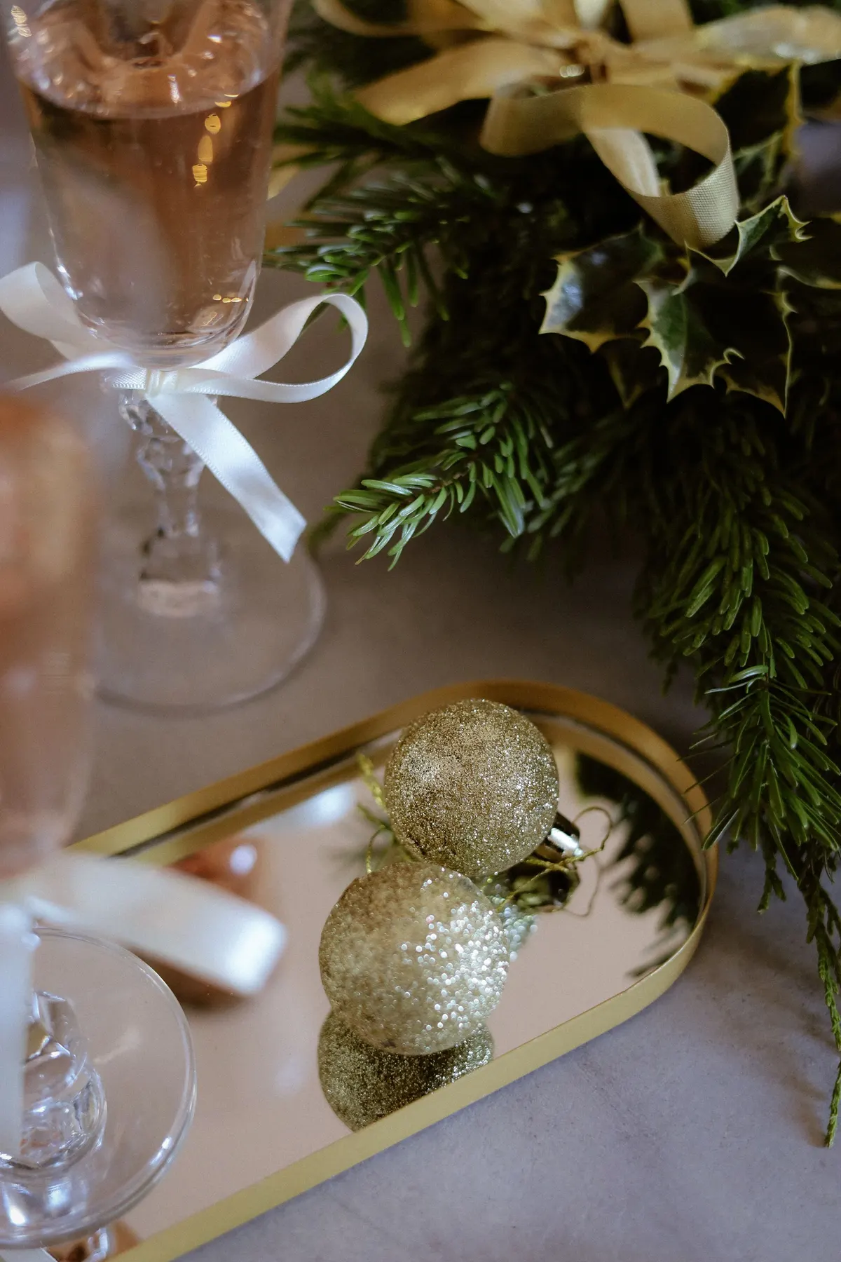 Gold-rimmed mirrored tray holding two gold glitter ornaments beside glasses, white ribbon bows and holly