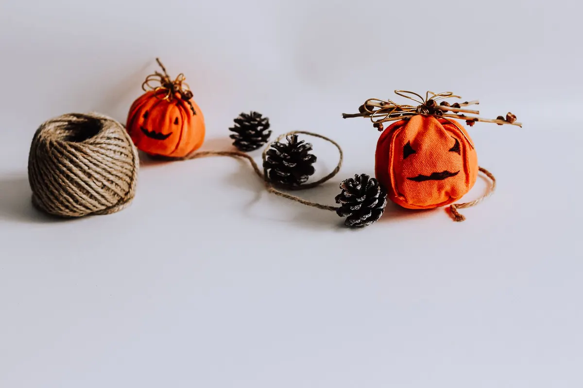 Two small orange fabric pumpkins with black felt jack-o'-lantern faces and twig stems beside twine and pine cones