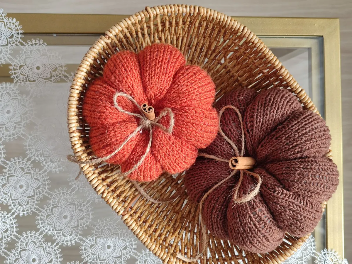 Coral and rust-brown knitted pumpkins with wooden stems and jute bows in a shallow wicker basket on lace