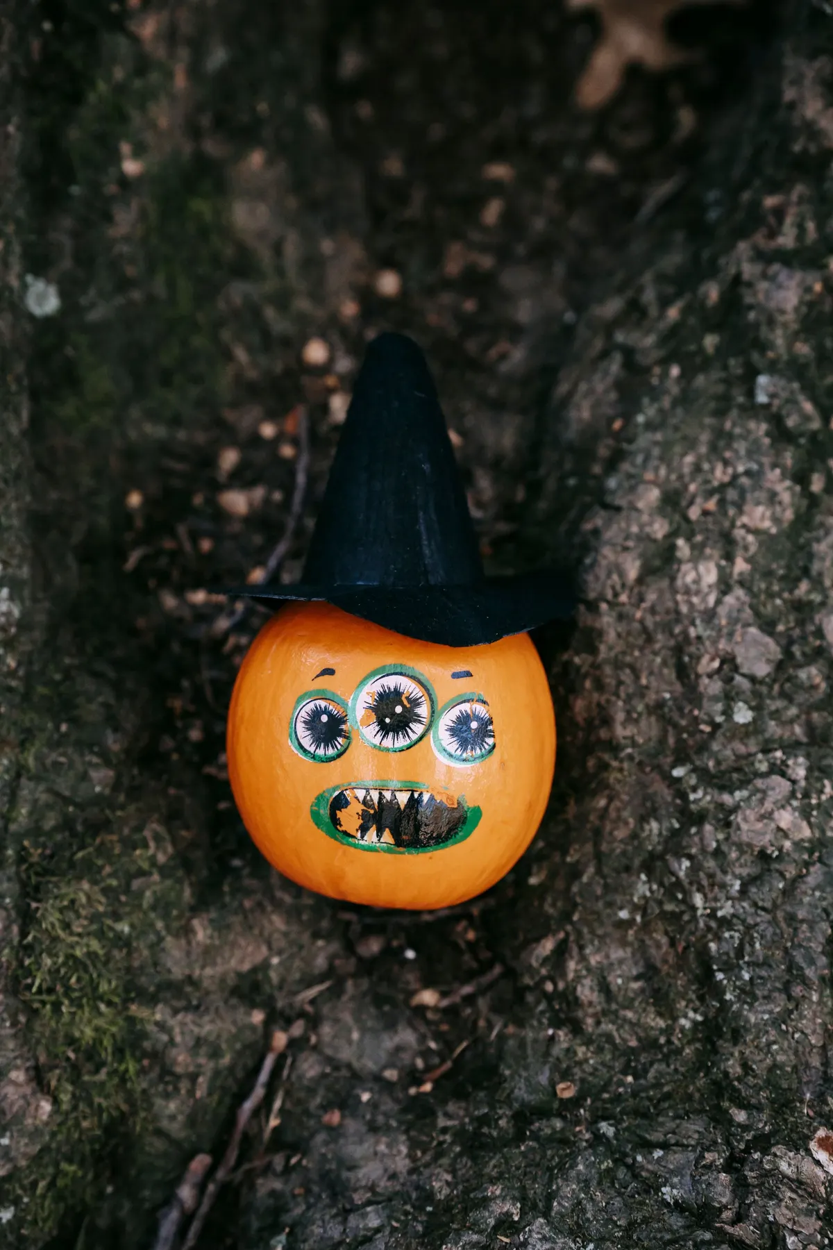 Small orange pumpkin painted as a three-eyed monster with jagged white teeth, wearing a black witch hat