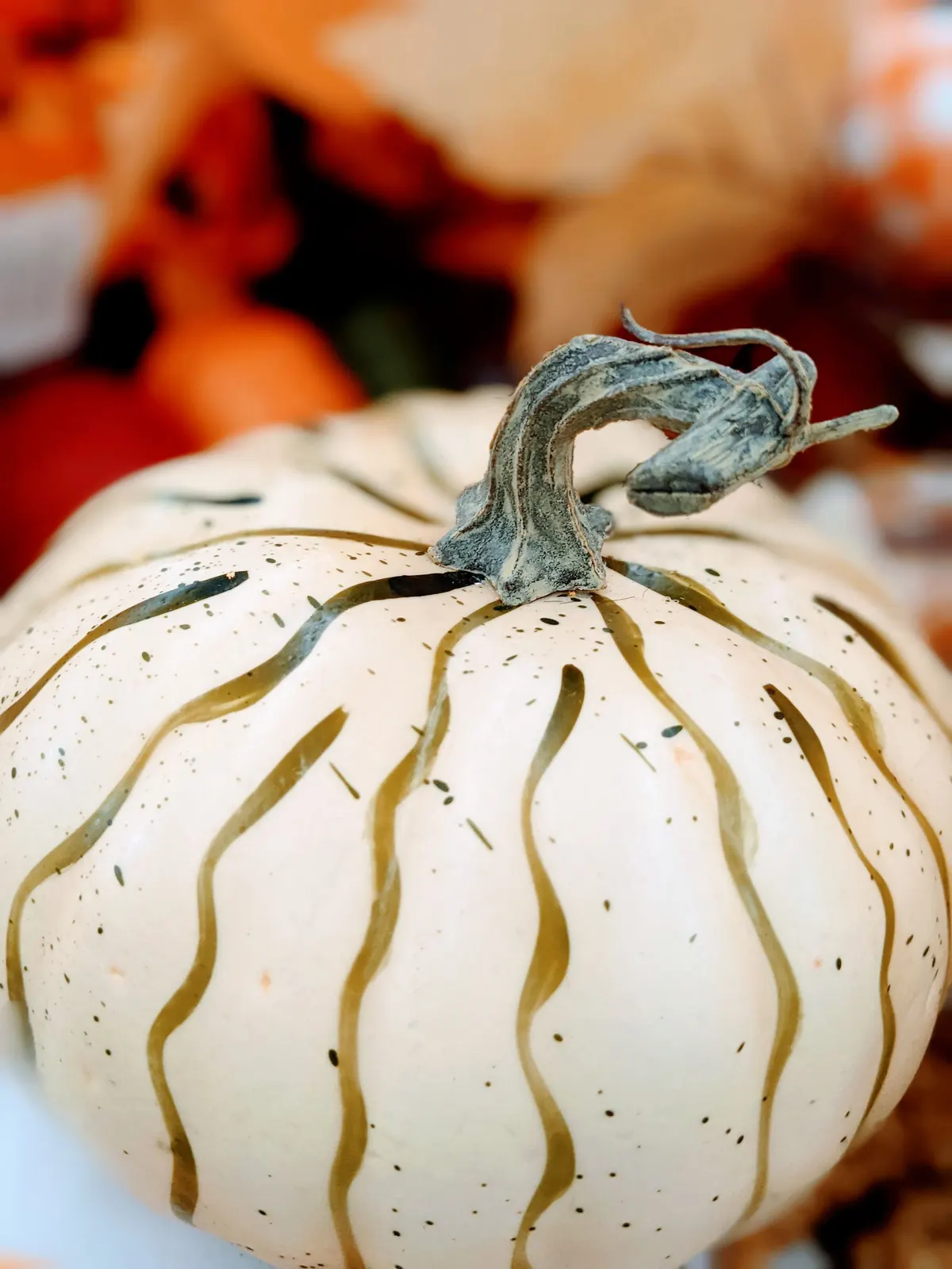 Cream-white pumpkin with freehand wavy olive-gold painted stripes and fine dark speckles, orange flowers blurred behind