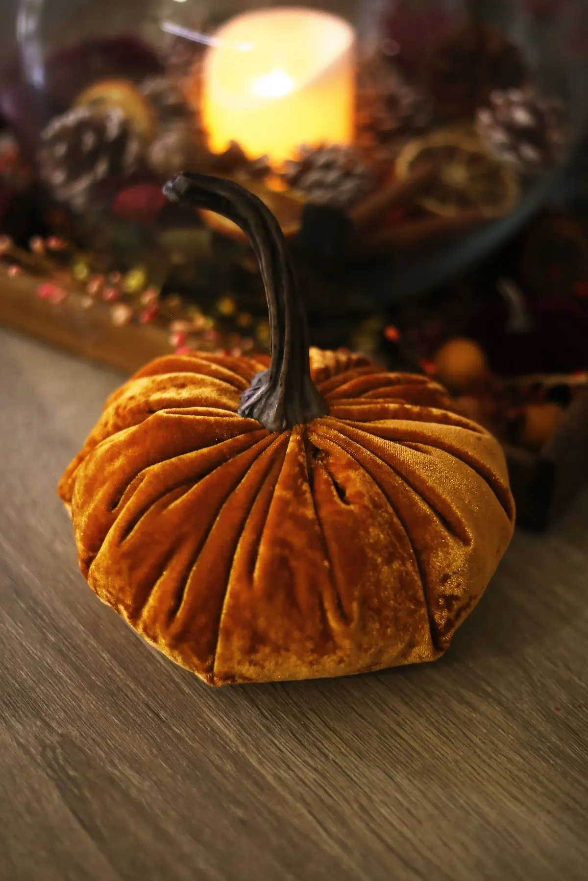 Burnt-orange crushed velvet pumpkin with a real dried stem on a wooden table, candle and pine cones behind