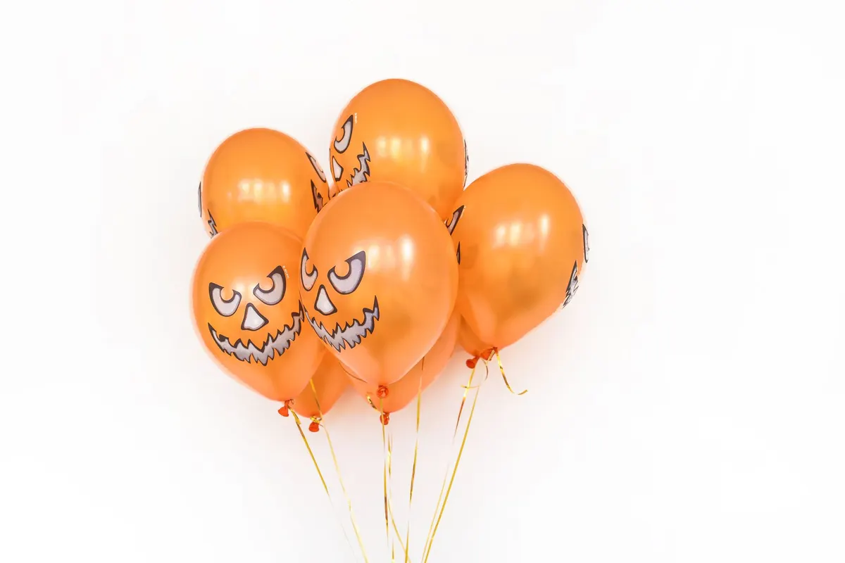Six or seven glossy orange balloons with black and white jack-o'-lantern faces tied to curling gold ribbons on white