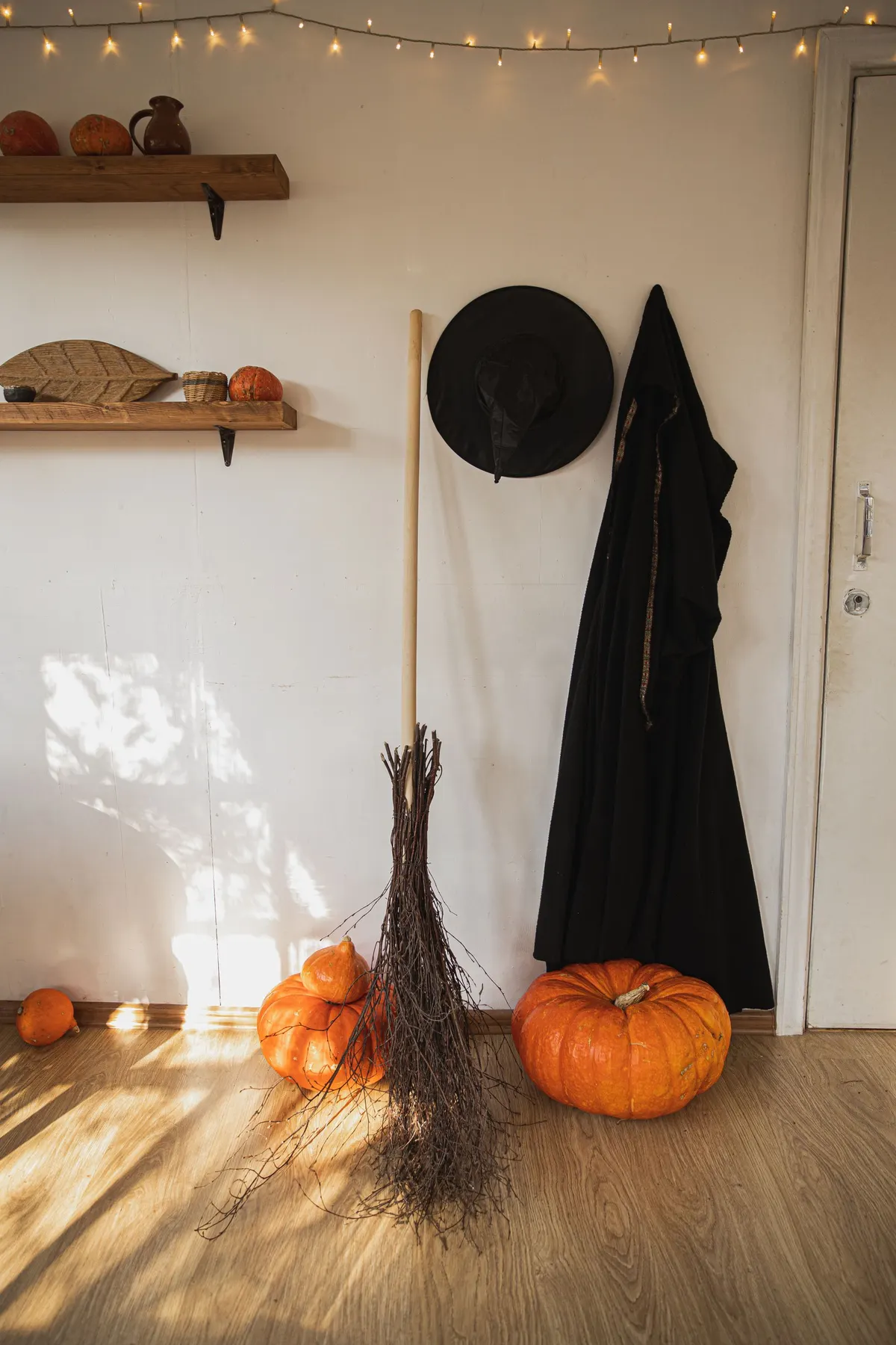 Black witch hat and long black cloak on wall hooks beside a twig broom, orange pumpkins