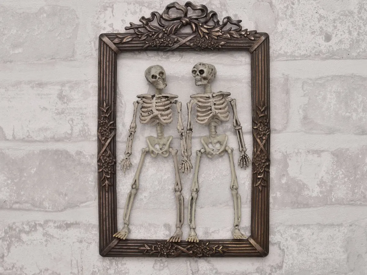 Two cream plastic skeletons standing side by side inside an ornate bronze picture frame on a whitewashed brick wall