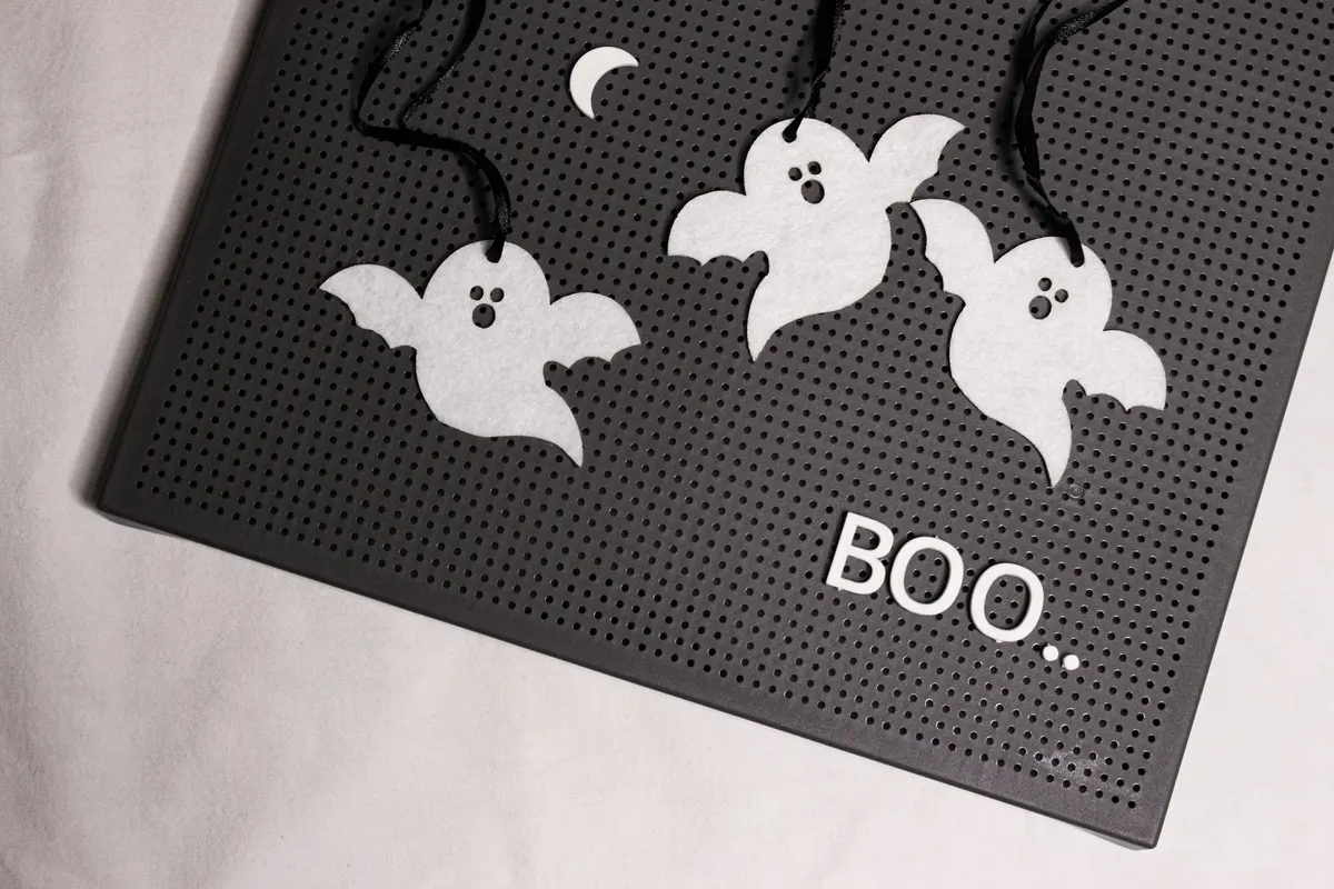 Black pegboard letter board tilted on a white cloth with three white felt ghosts