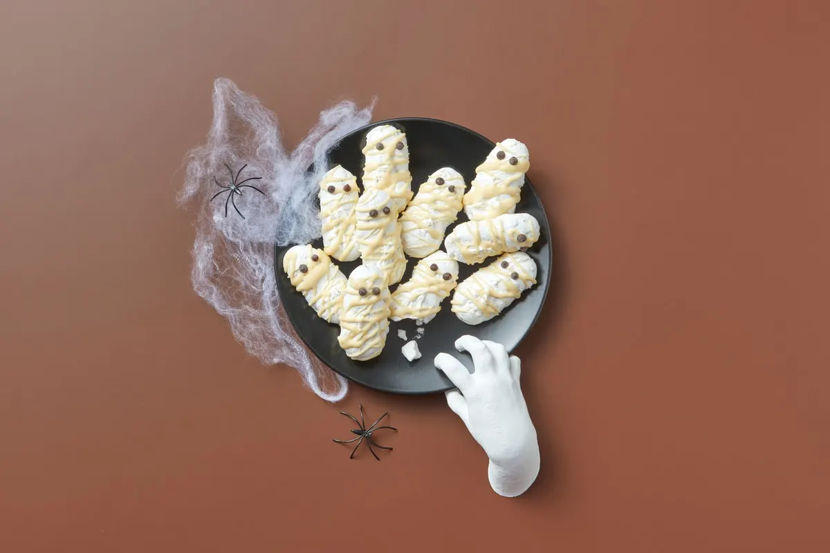 Round black plate of about ten white-iced mummy cookies with candy eyes, beside stretch cobweb and plastic spiders