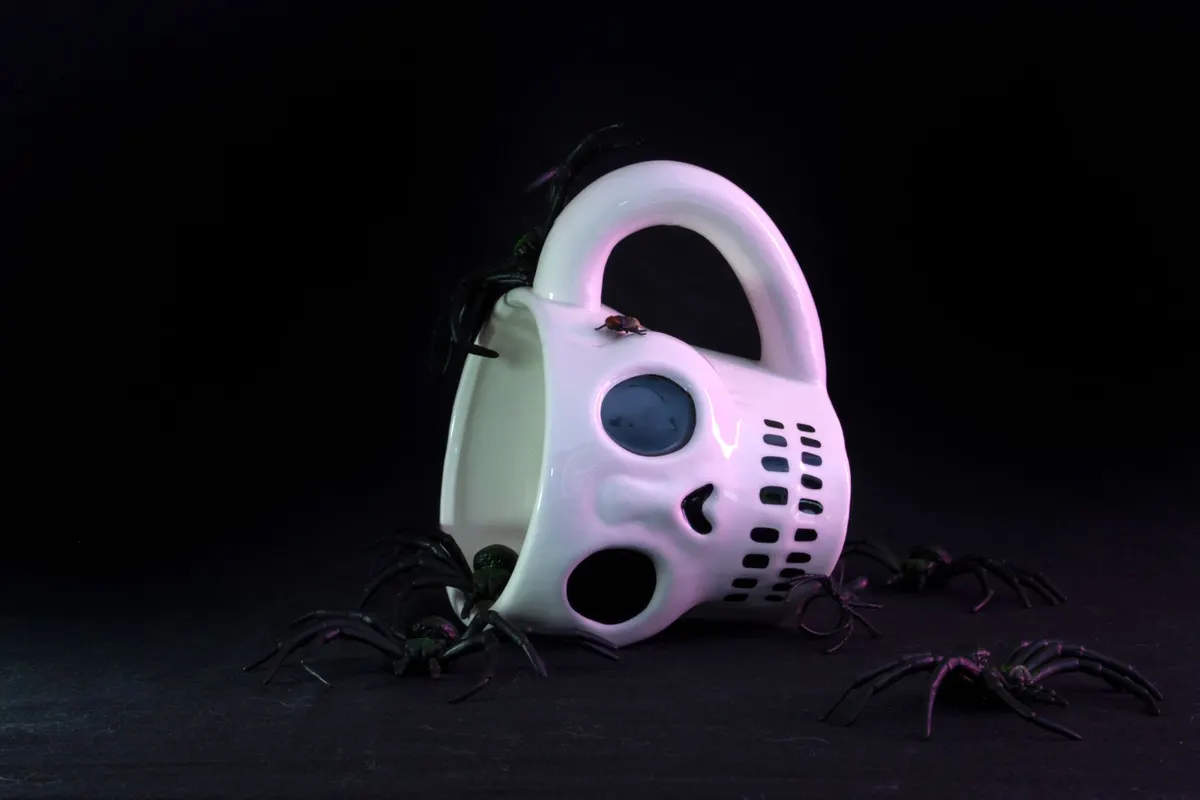 White glossy ceramic skull mug lying on its side with five or six black plastic spiders on and around it
