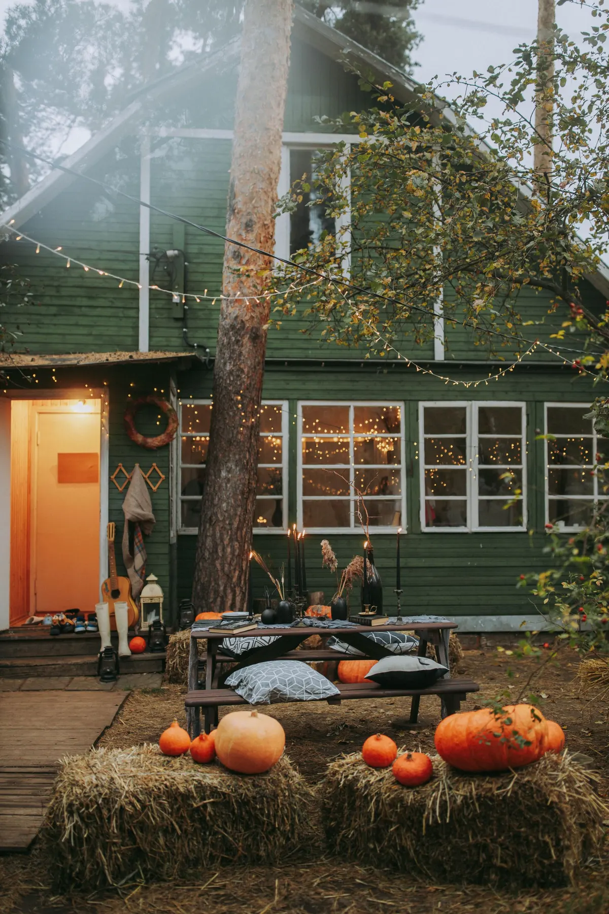 Backyard at dusk with a wooden picnic table and cushions, hay bales, about eight pumpkins and string lights