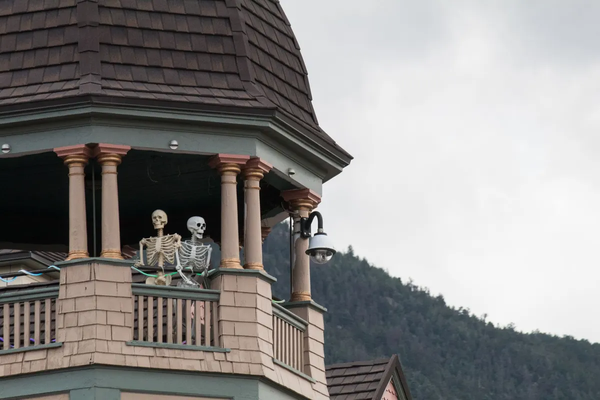 Two ivory skeletons posed on a Victorian-style wooden balcony under a shingled tower with wooded hills in the background