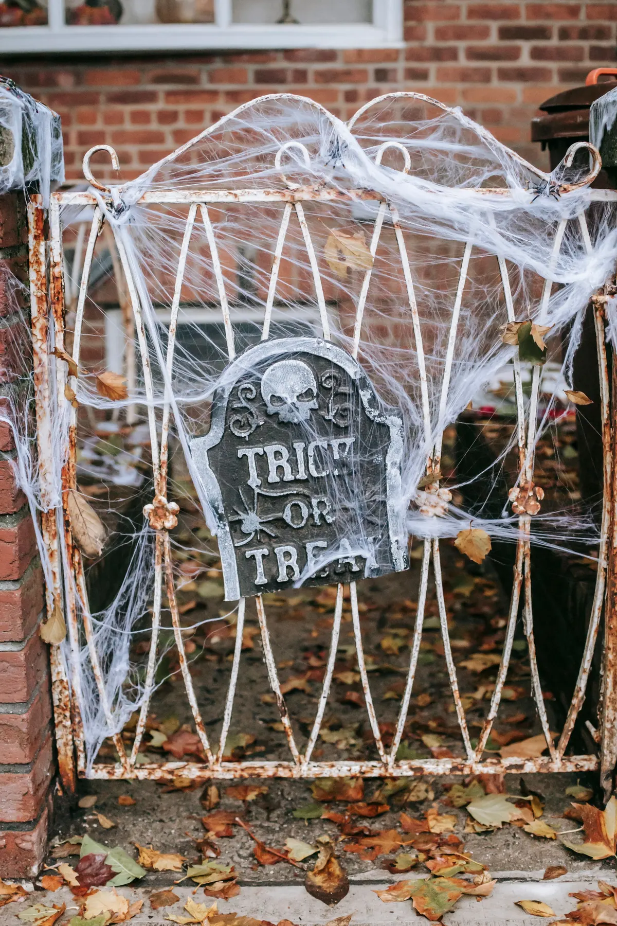 Gray foam tombstone with a skull tied to a white iron gate and covered in cobwebs, with fallen leaves behind