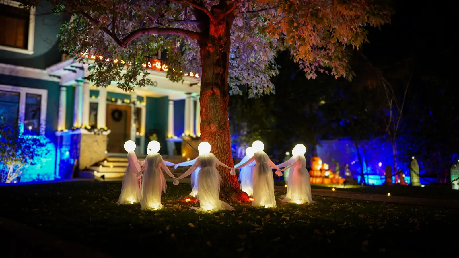 Glowing white ghosts with round lit heads holding hands in a ring around a large tree at night