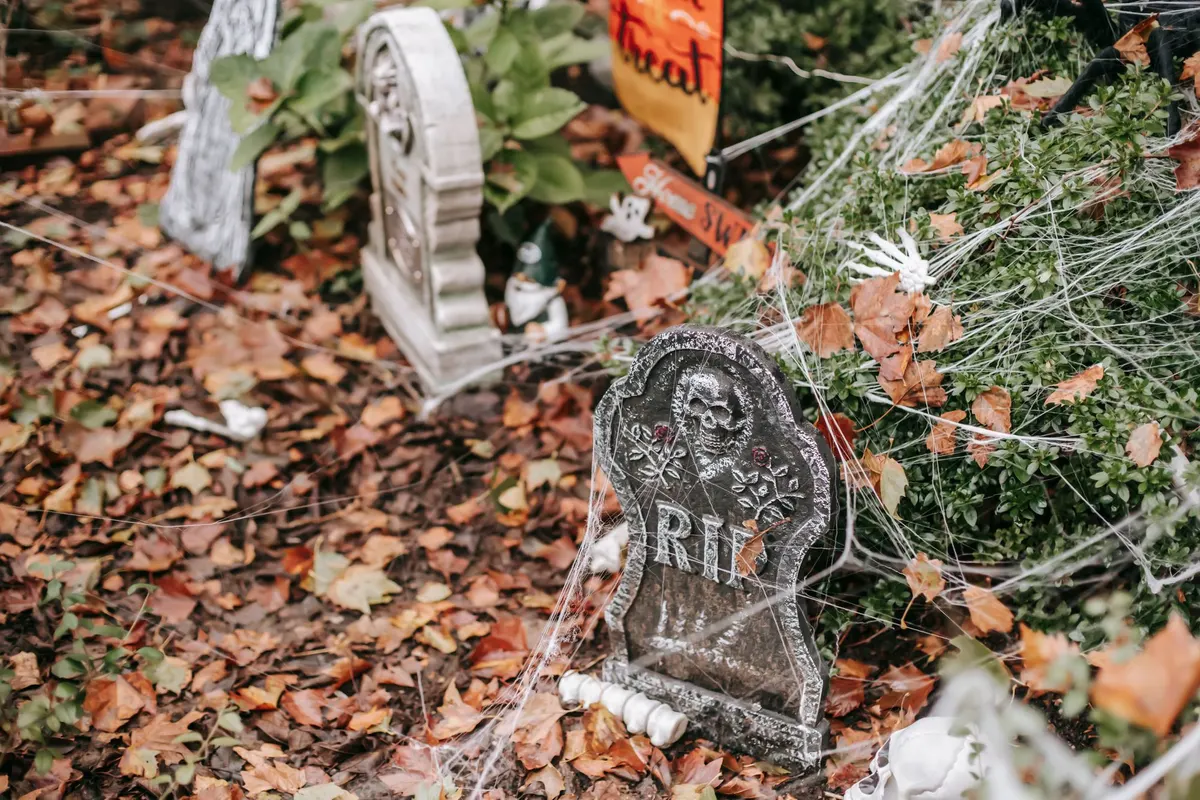 Three gray foam gravestones, one marked RIP, wrapped in white cobwebs among brown leaves with skeleton hands and orange signs