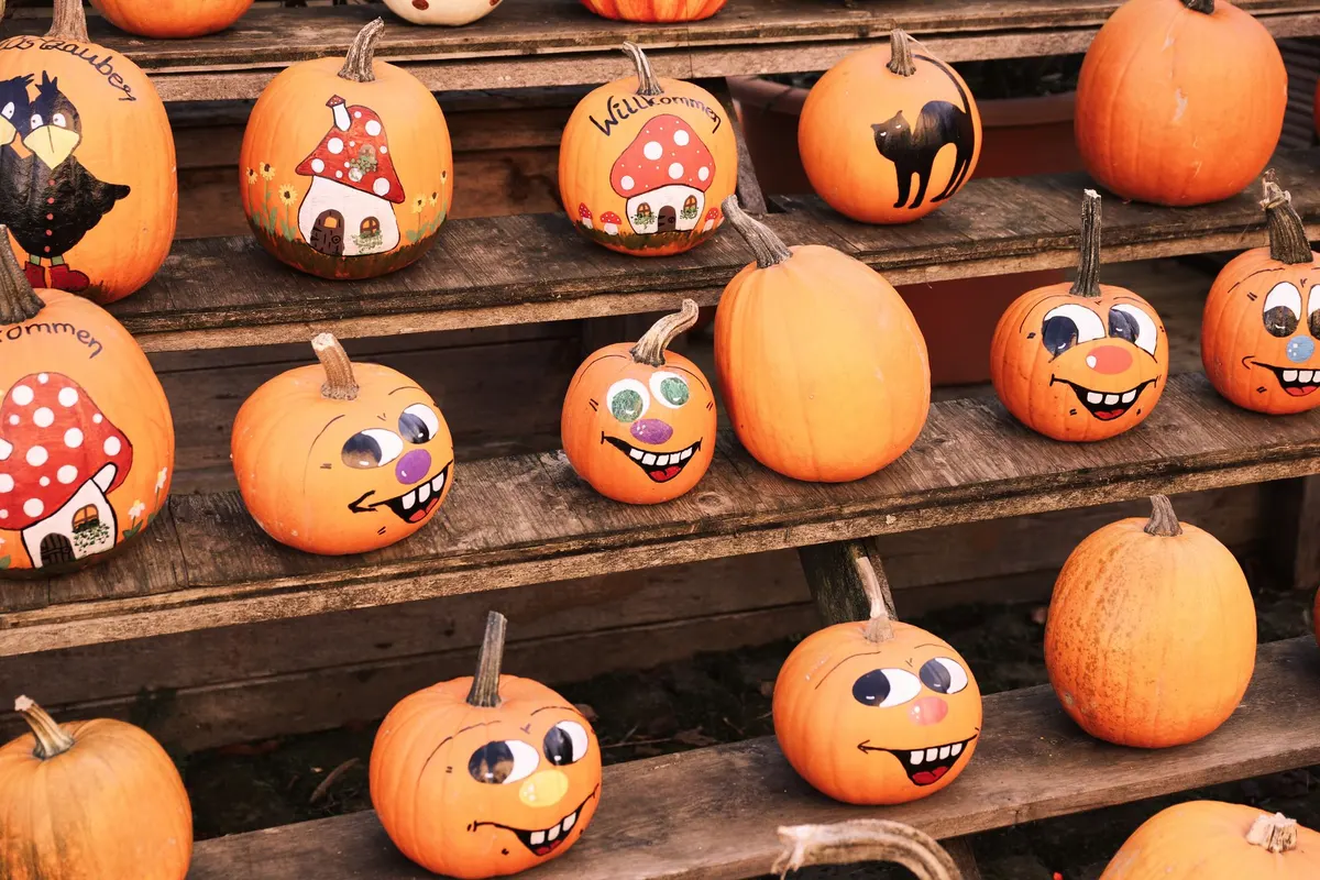 Orange pumpkins on weathered wooden benches painted with grinning cartoon faces, mushroom houses and a black cat, some left plain