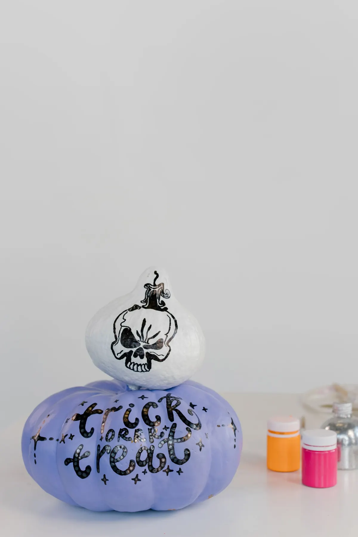 Small white pumpkin painted with a black skull sits on a lavender pumpkin lettered trick or treat with silver speckles
