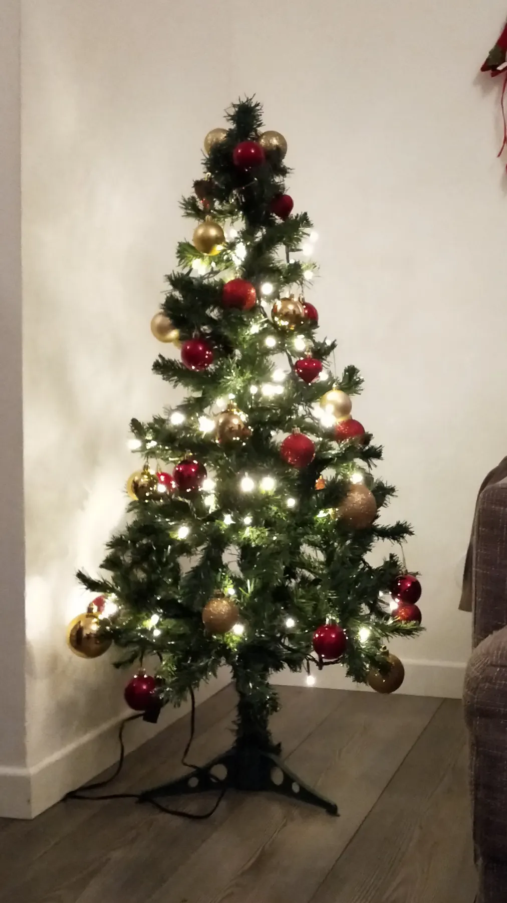Small pre-lit artificial Christmas tree with red and gold baubles standing in a corner of a living room