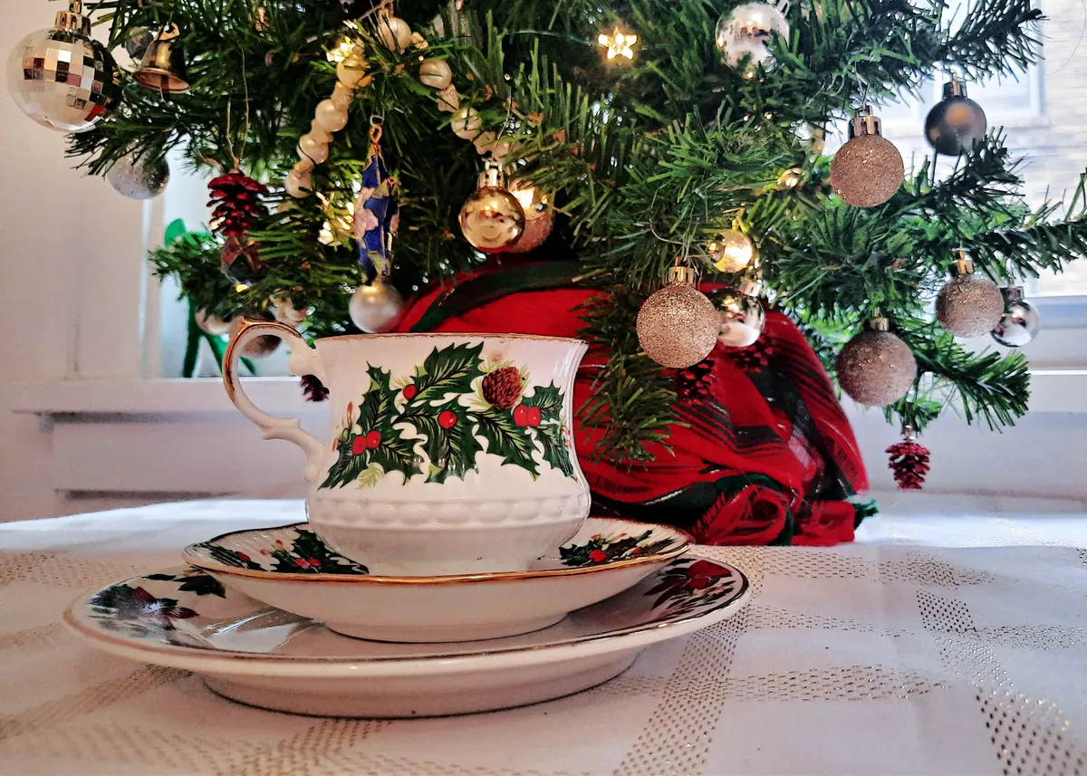 Small tabletop Christmas tree with fairy lights and a plaid skirt behind a holly-pattern teacup